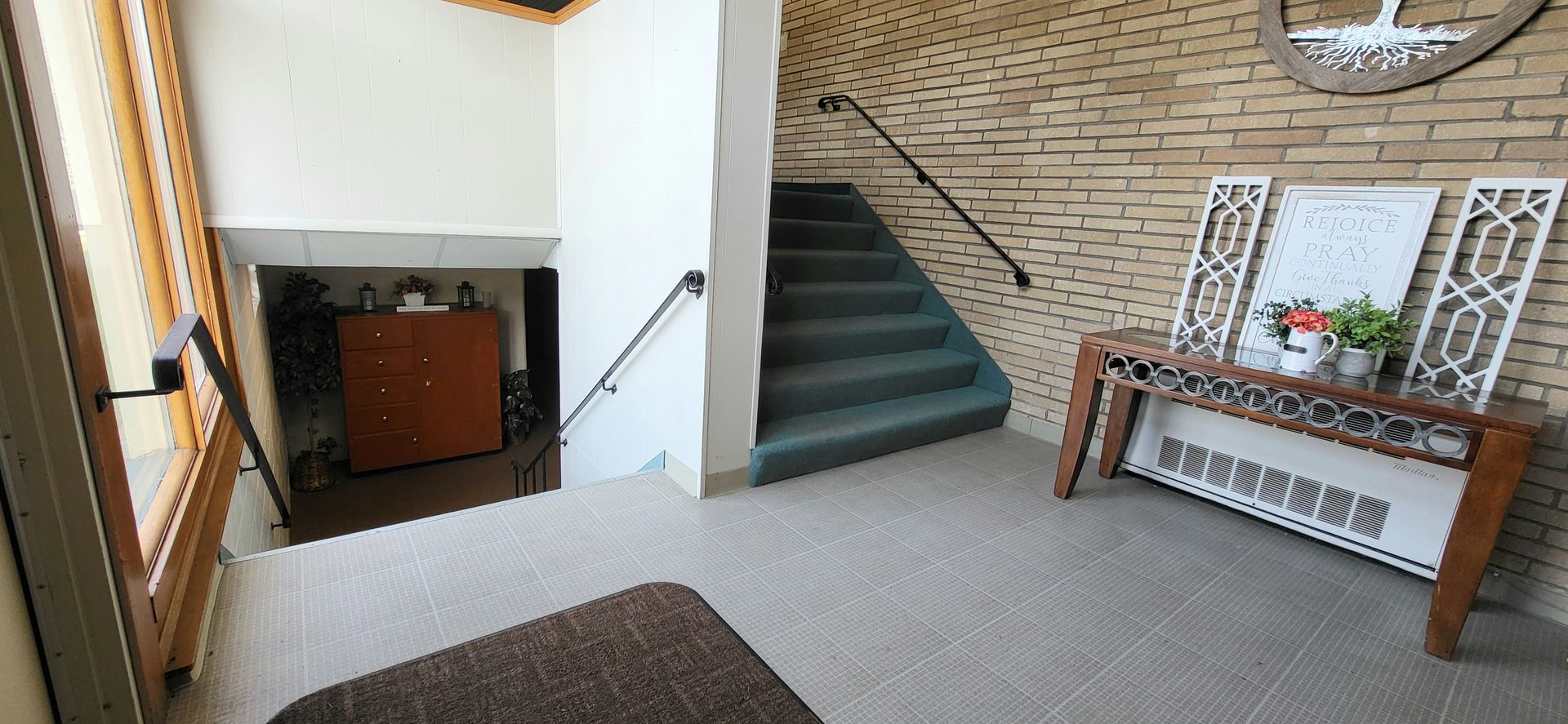 A building entryway featuring a staircase with green carpet, a wooden table with decor against a brick wall, and a lower level visible.