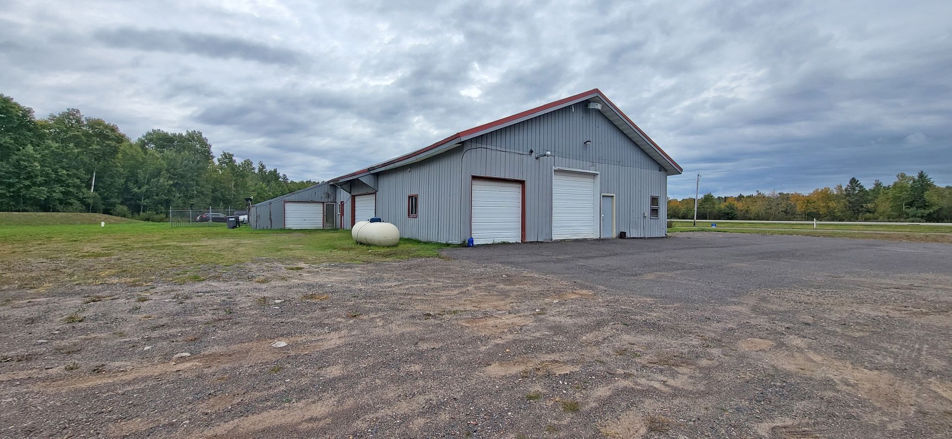 Gray metal barn on a gravel lot under cloudy skies, with trees and open field in the background