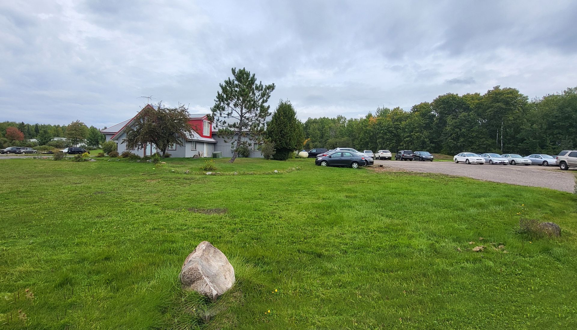 Grassy field with a large rock in front, trees and parked cars under a cloudy sky