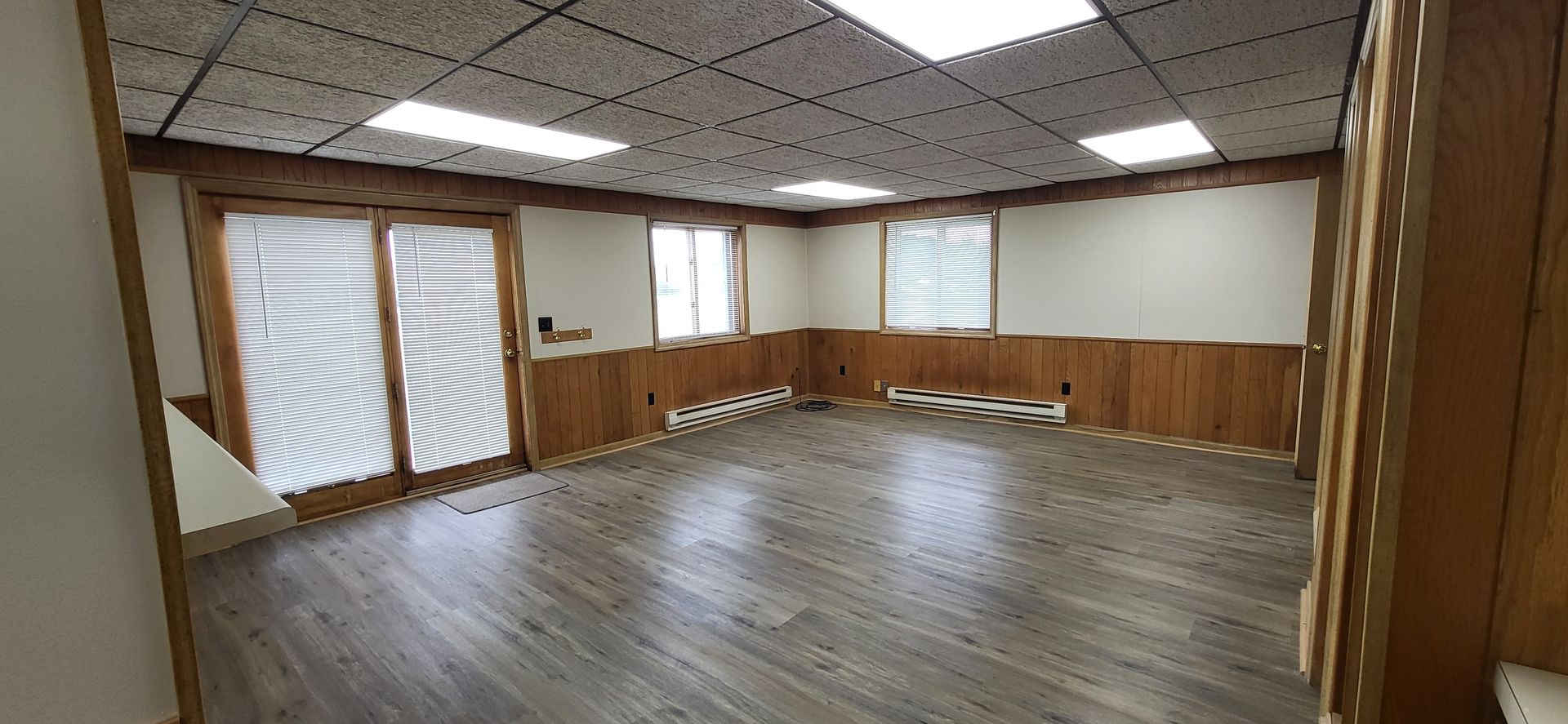 Empty wood-paneled room with gray floor, white ceiling tiles, and windows with curved blinds on one wall