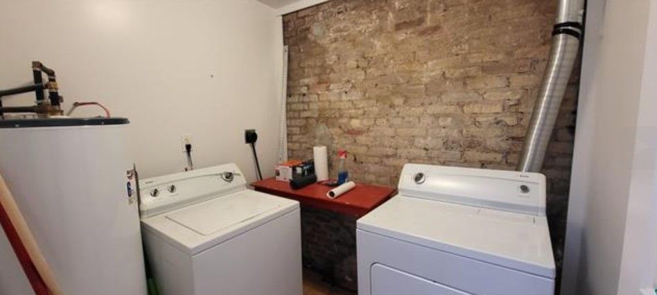 Laundry room with two white top-loading washing machines, a red shelf, and an exposed brick wall.