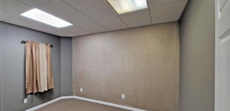 Empty beige room with gray walls, carpeted floor, window curtain, and fluorescent ceiling lights