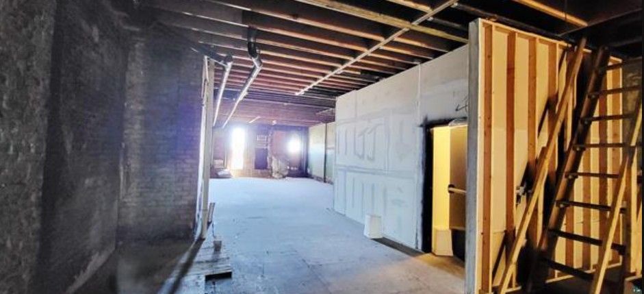 Dim unfinished hallway with exposed beams, drywall, a ladder, and bright light at the far end.