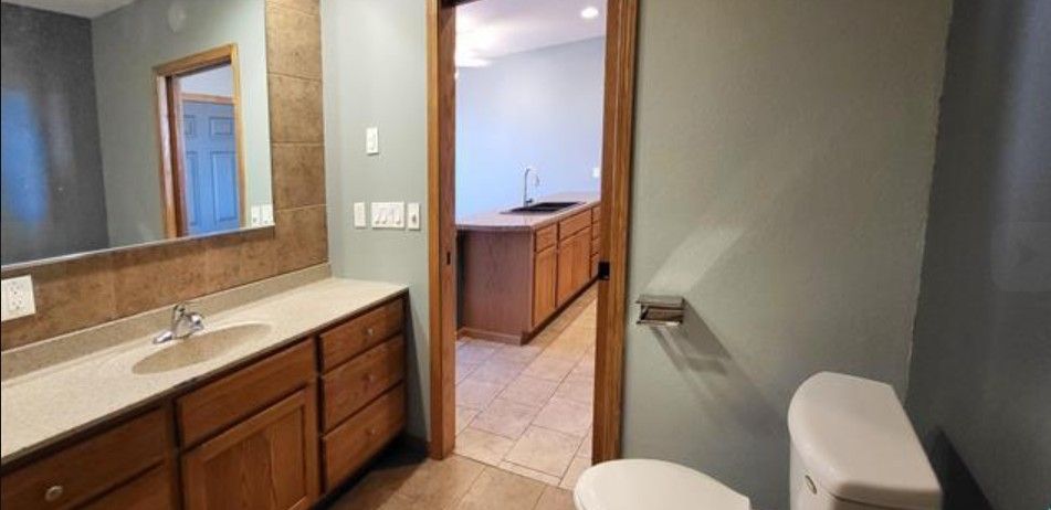 Bathroom vanity and toilet beside an open doorway into a kitchen with wood cabinets and a granite counter.