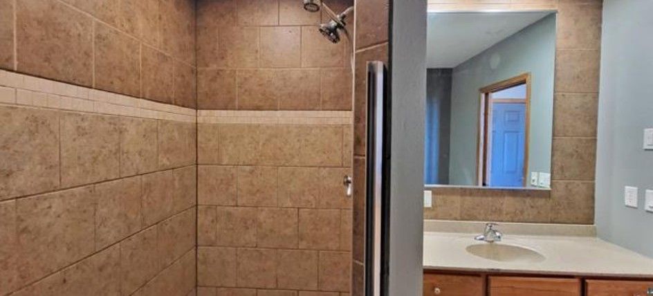 Bathroom with tiled shower and vanity sink, seen through an open glass shower door.