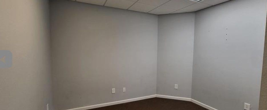 Empty gray office room with white trim, dark carpet, and electrical outlets on the walls