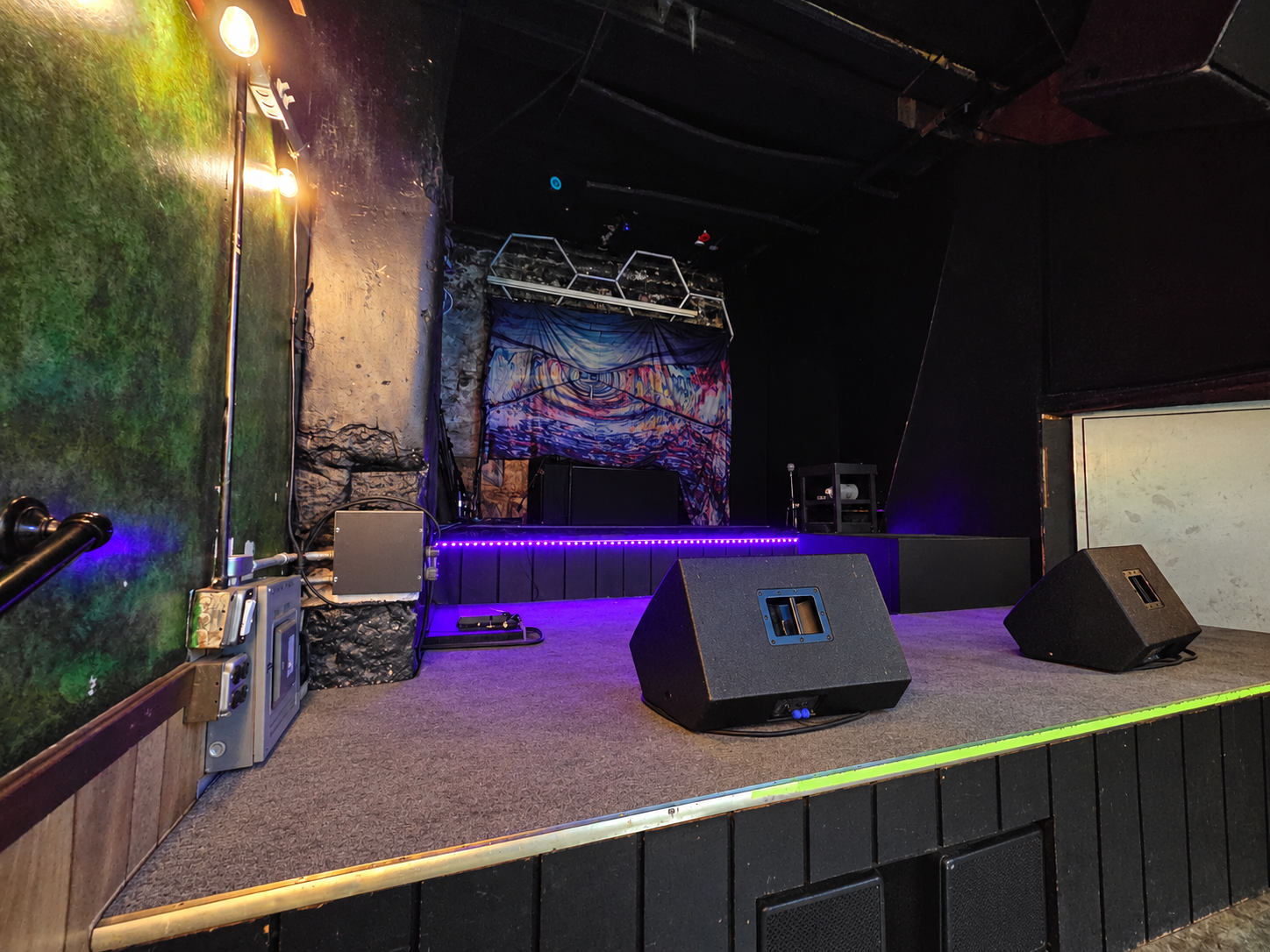 Empty small stage with purple lights, two floor speakers, a mural backdrop, and a white screen on the right