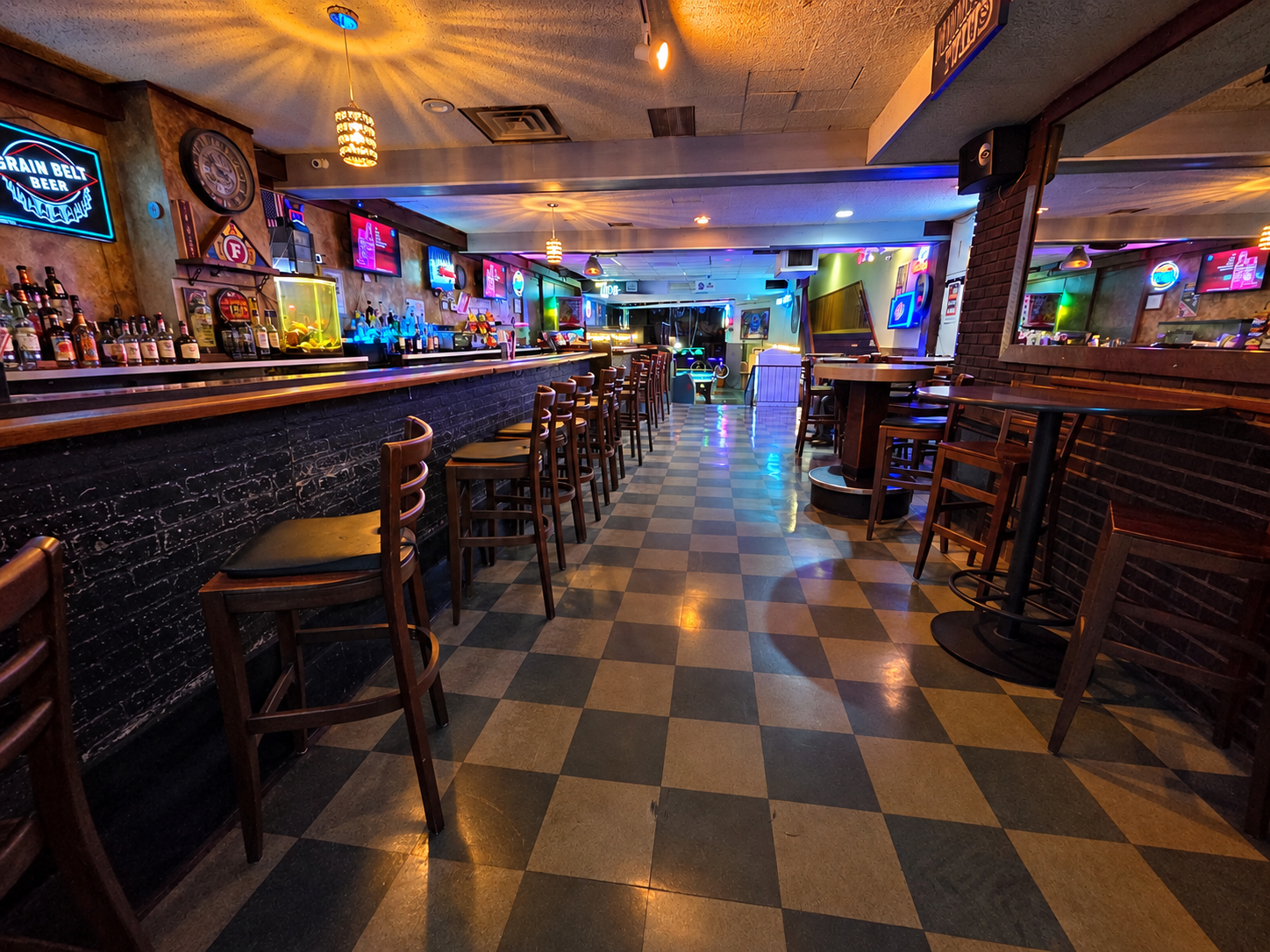 Dimly lit bar interior with checkered floor, wooden stools, and colorful neon lights behind the counter