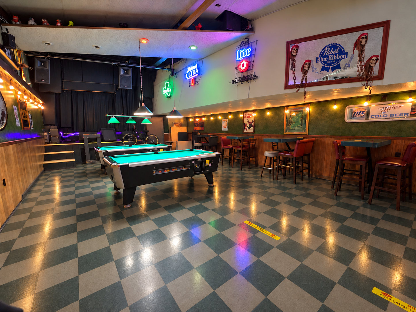 Dimly lit pool hall with green felt tables, checkered floor, bar stools, and neon signs