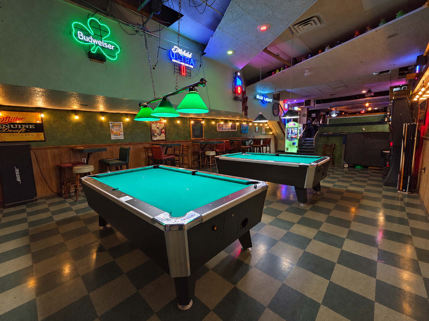 Neon-lit pool hall with two pool tables, checkered floor, and bar seating in the background
