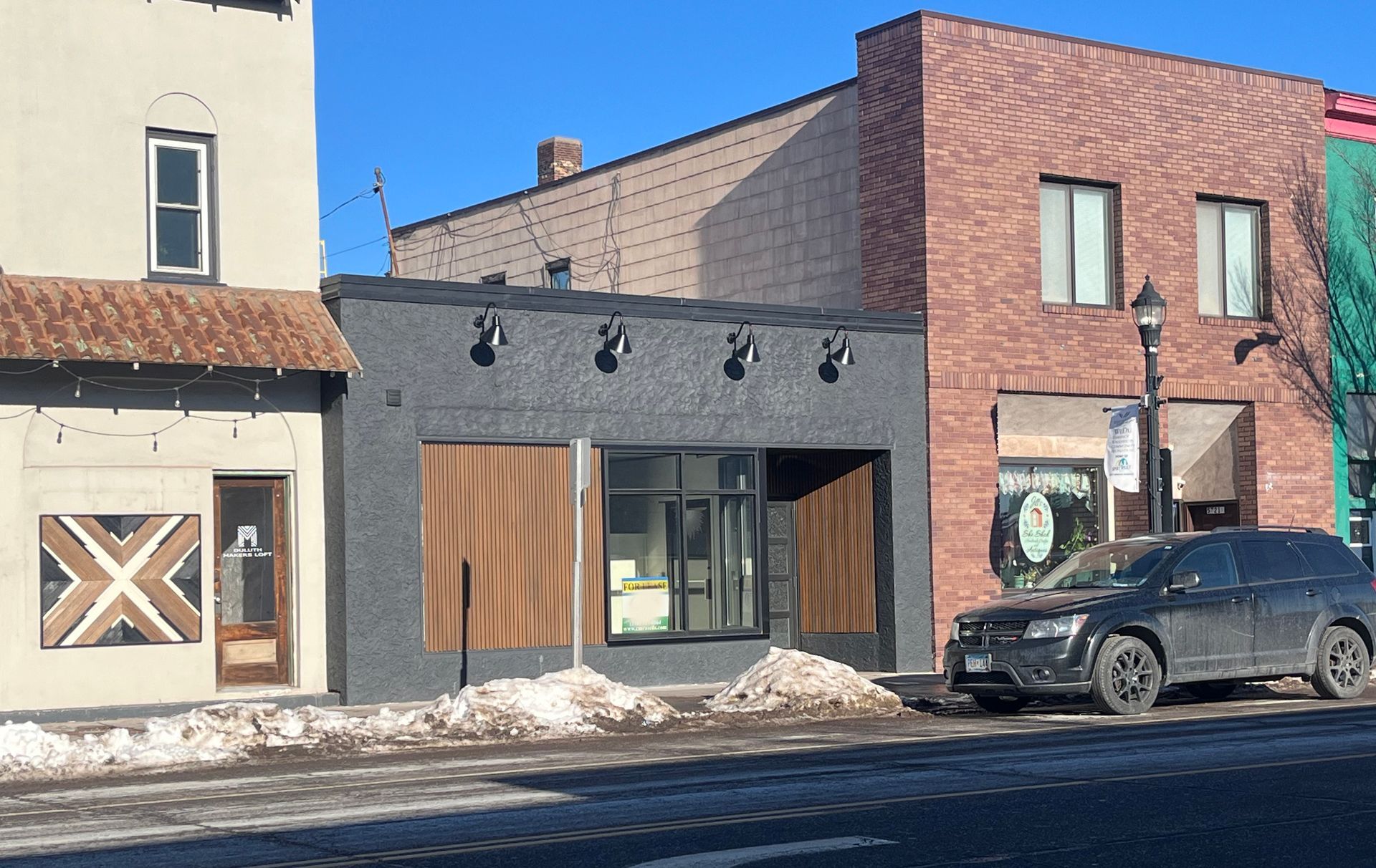 Buildings on a street, some with boarded-up windows. A car is parked on the street. Blue sky.