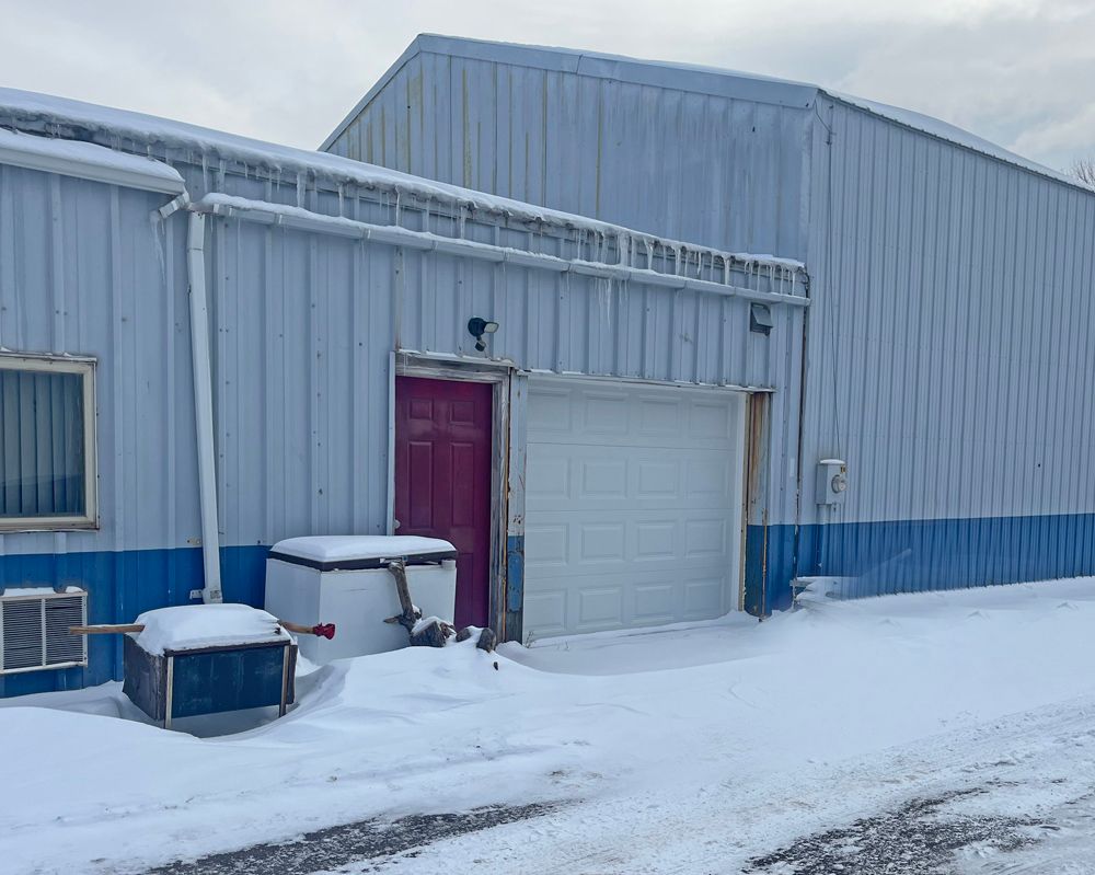Snow-covered industrial building with a pink door and a closed white garage door. Blue and white siding.