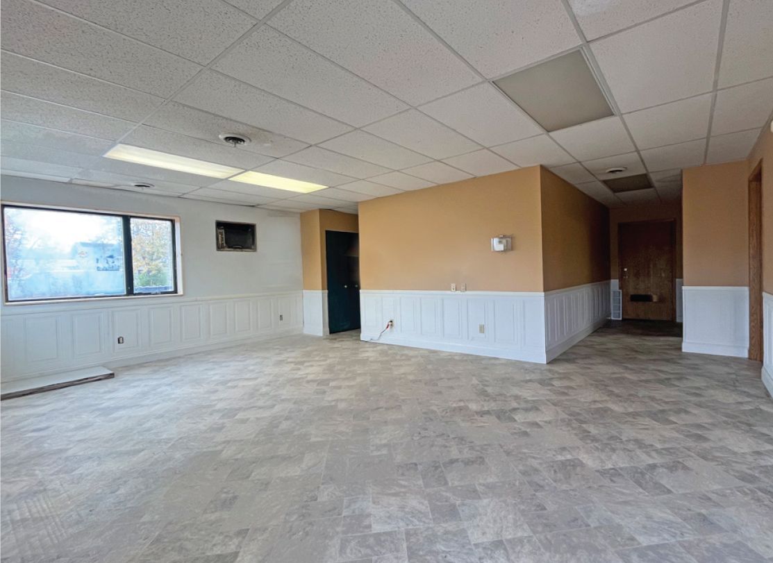 Empty commercial space with light-colored flooring, beige walls, and a dropped ceiling with fluorescent lights.