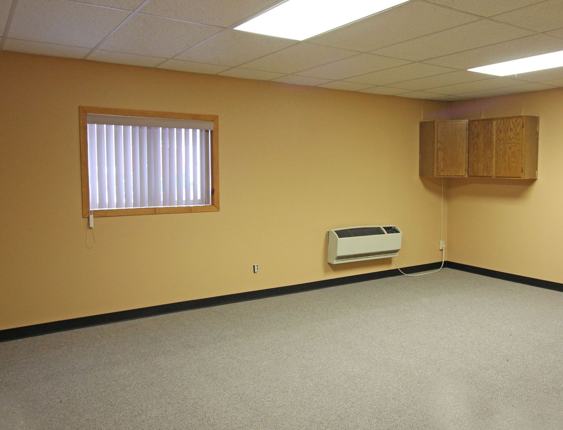 Empty room with beige walls, window, air unit, cabinets, and gray carpet.
