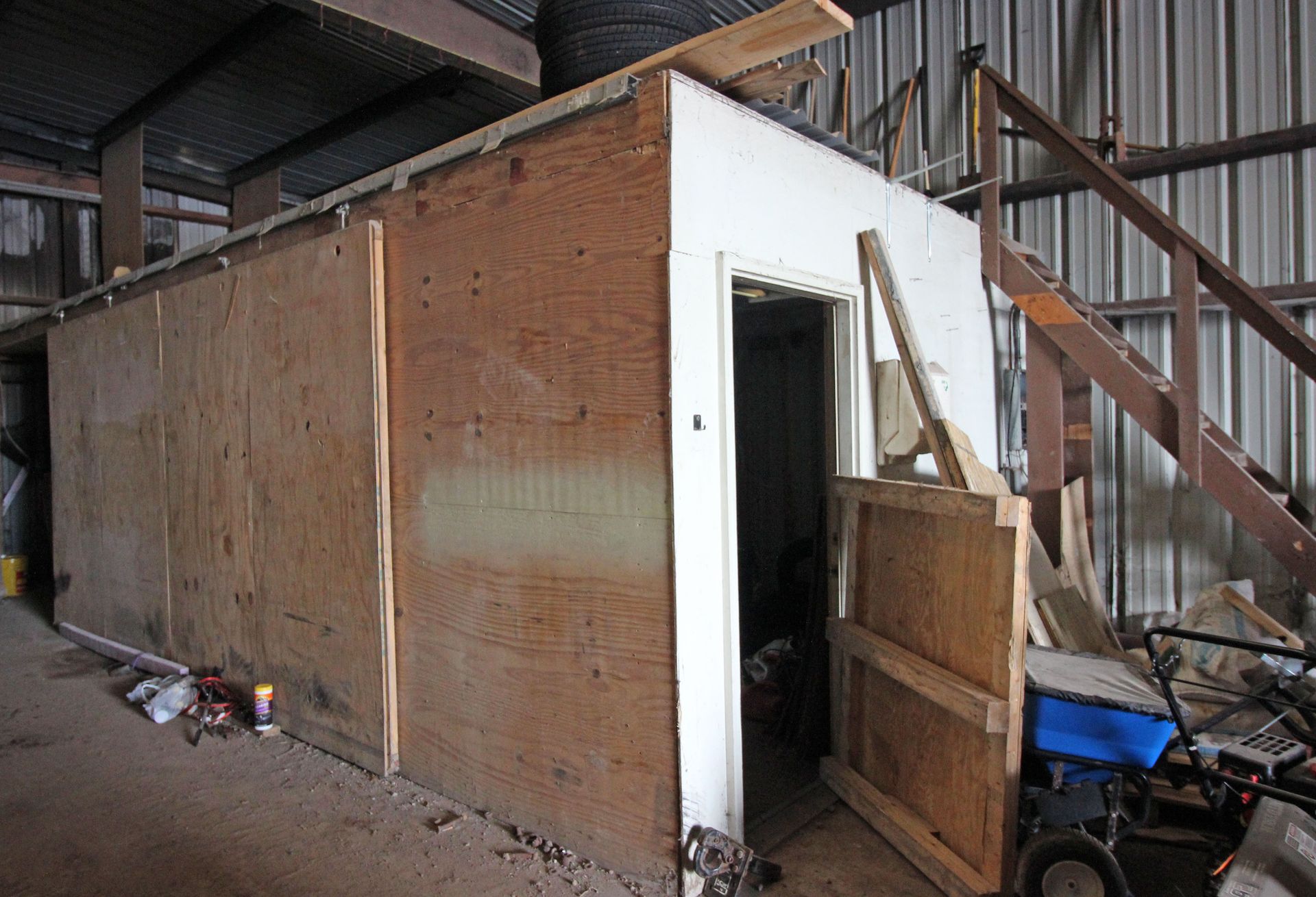 A plywood structure with a door inside a corrugated metal building. A wooden ramp sits nearby.