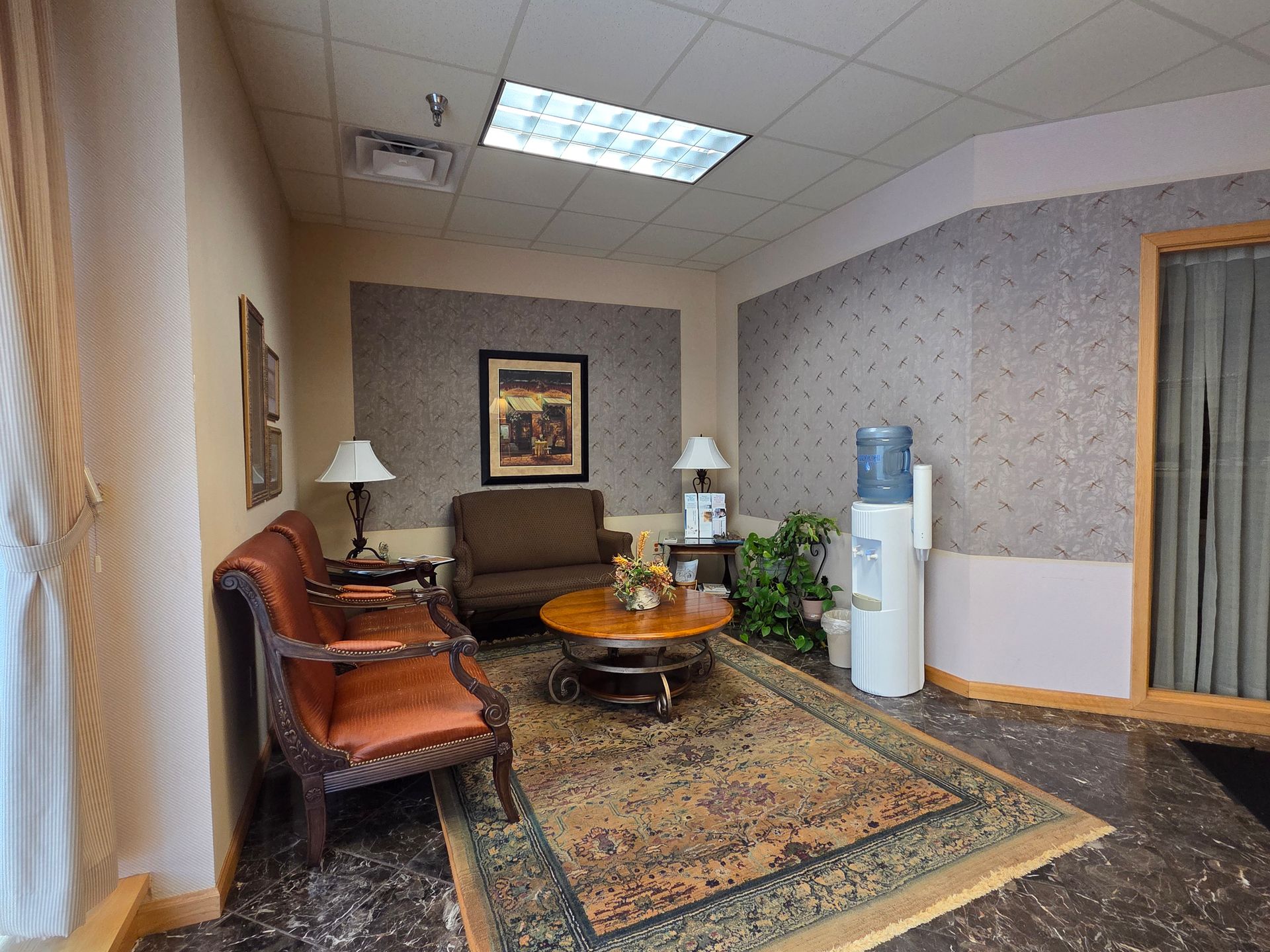 Waiting room: chairs, rug, and water dispenser against textured walls, art, and a round table.