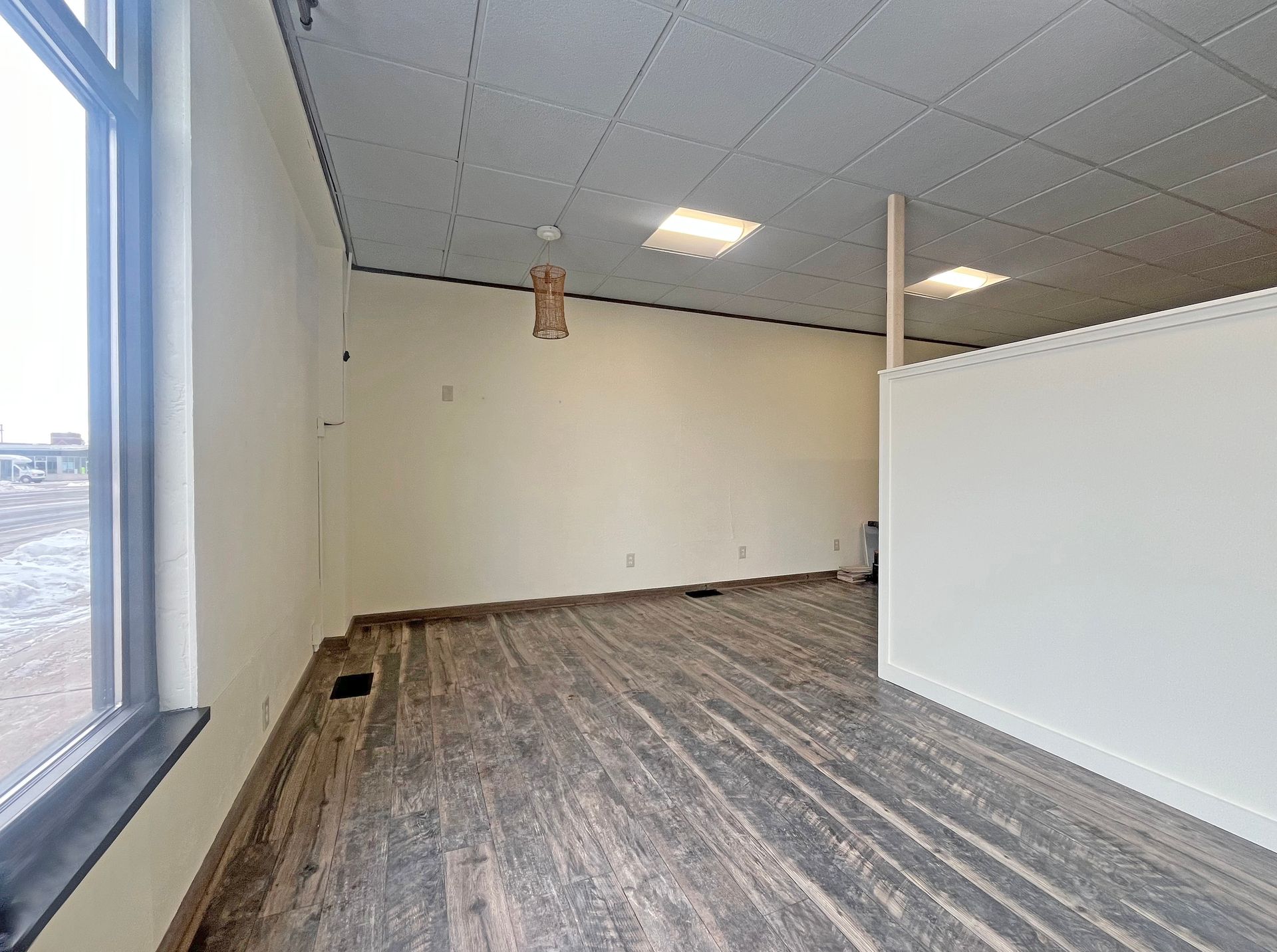 Empty commercial space with large window, neutral walls, and wood-look flooring.