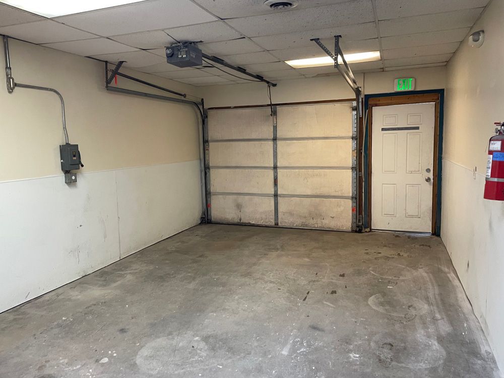 Empty garage with open door, exit sign, fire extinguisher. Concrete floor, beige walls, overhead door opener.