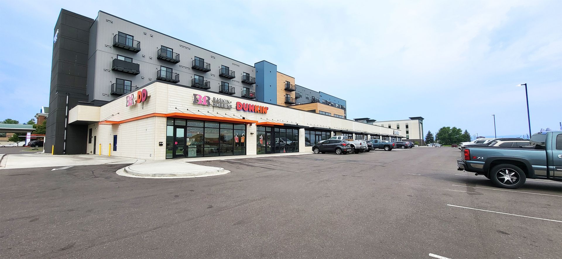 A strip mall with several businesses under a multi-story building; parking lot in the foreground; blue sky overhead.