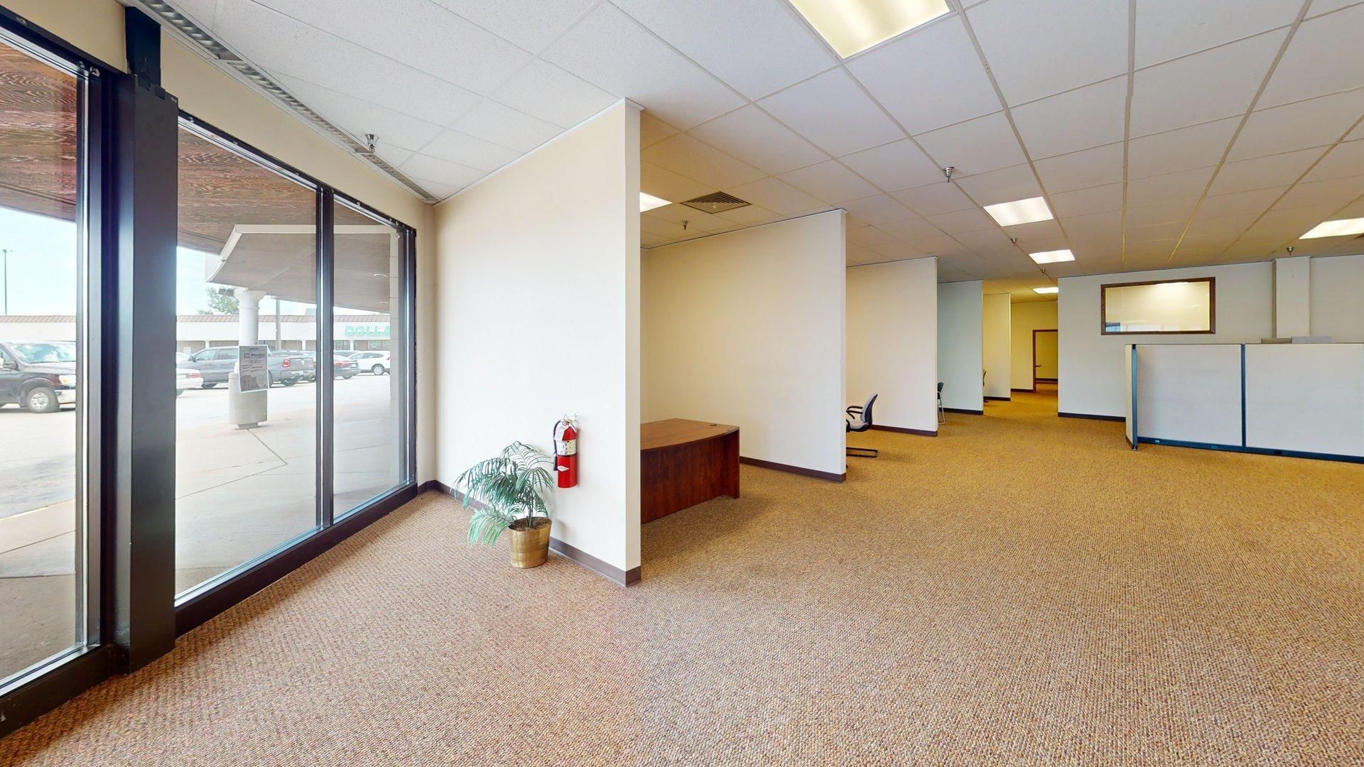 Empty office with carpet, large windows, and cubicle dividers. Brown desk and beige walls.