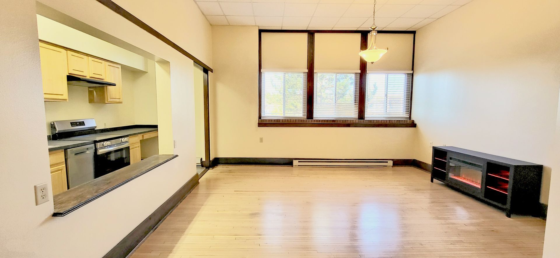 Empty living room with wood floor, white walls, kitchen pass-through, and window blinds