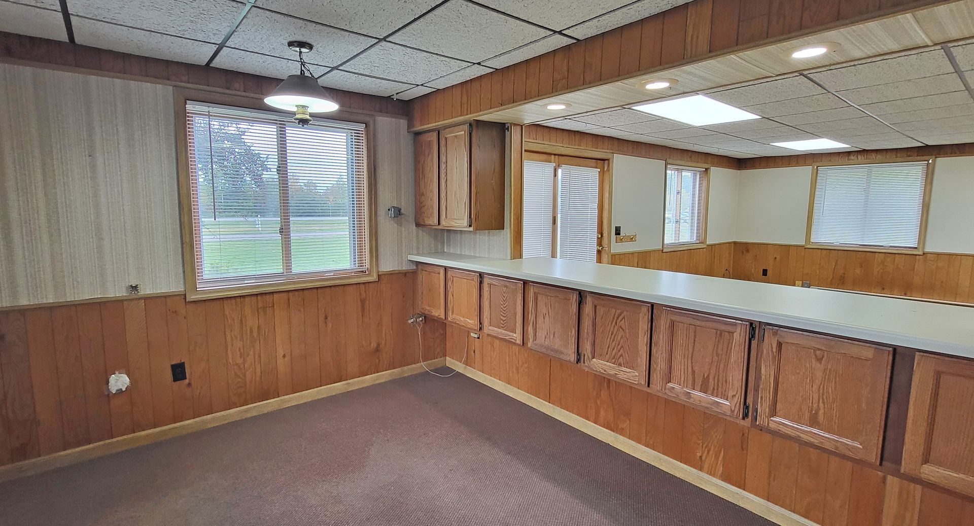 Empty wood-paneled room with carpet, window, and long mirrored counter under fluorescent lights
