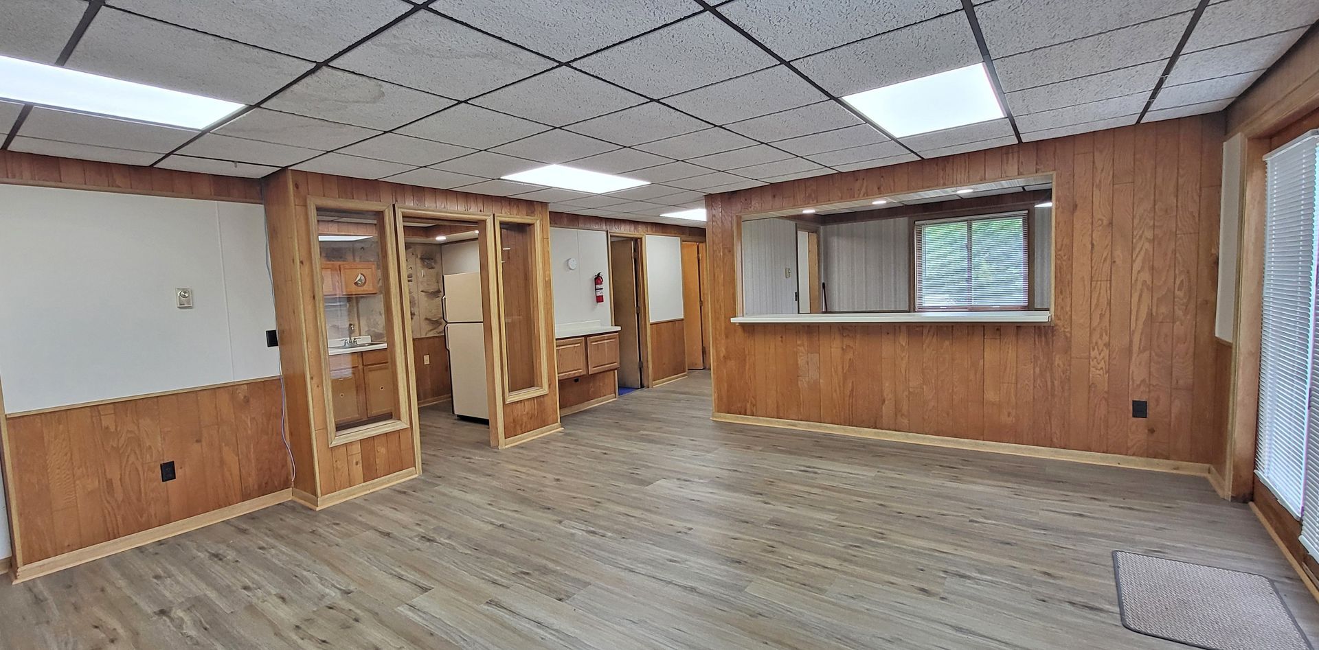 Empty wood-paneled room with tiled ceiling, large window, and open doorway.