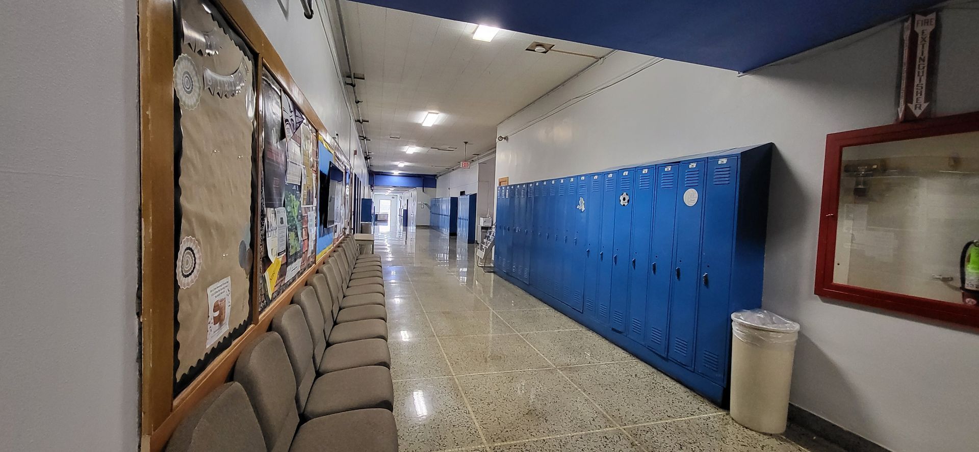 School hallway with blue lockers on the right, bulletin boards and seating on the left, and a trash can.