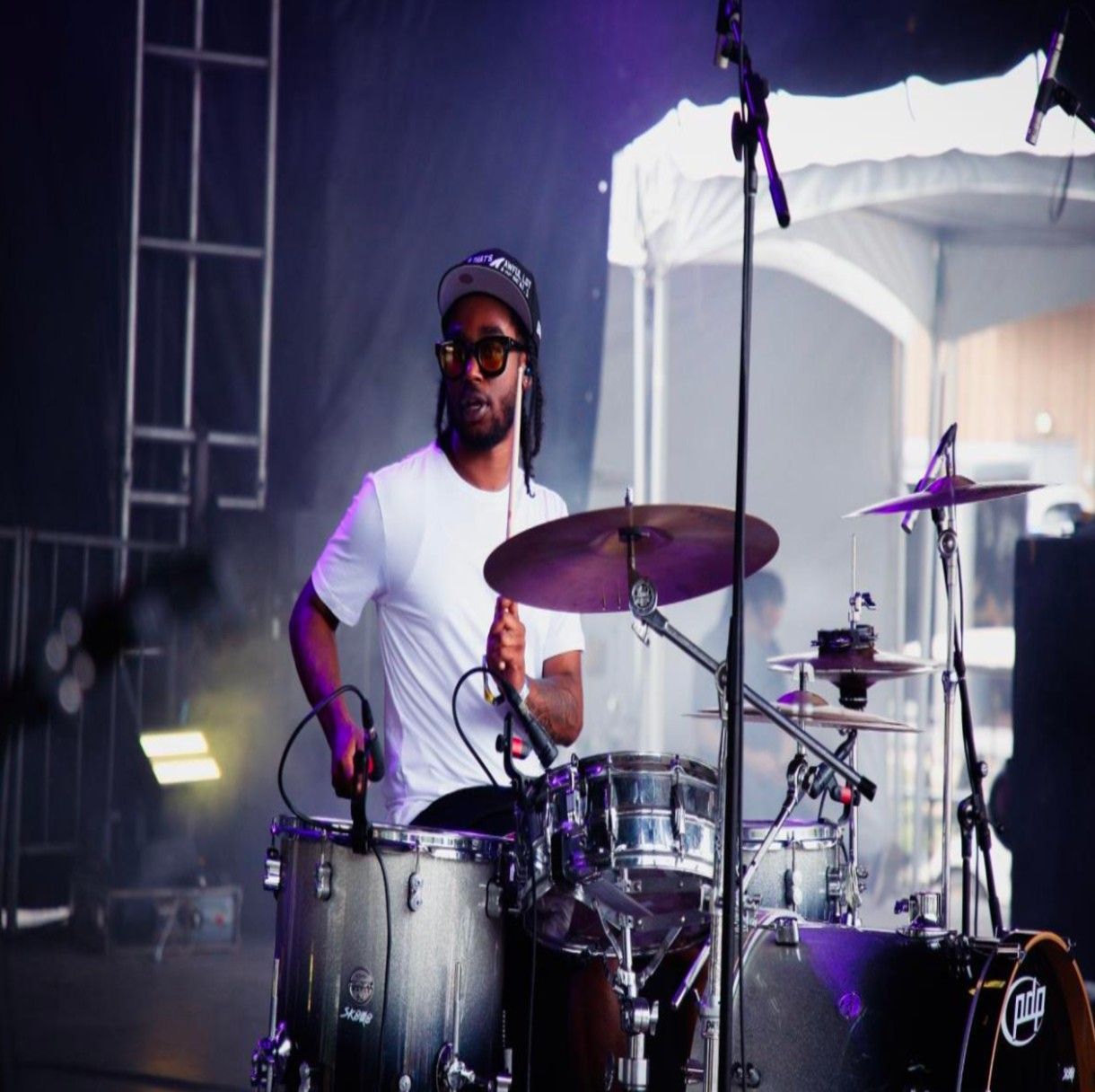 Drummer in white shirt plays on stage, dark skin, wearing sunglasses, drum set visible.