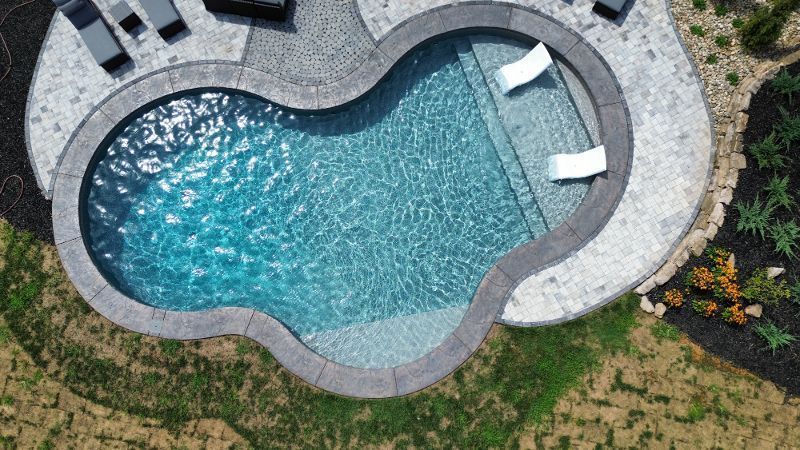 Overhead view of a kidney-shaped pool with two white lounge chairs inside and gray stone patio.