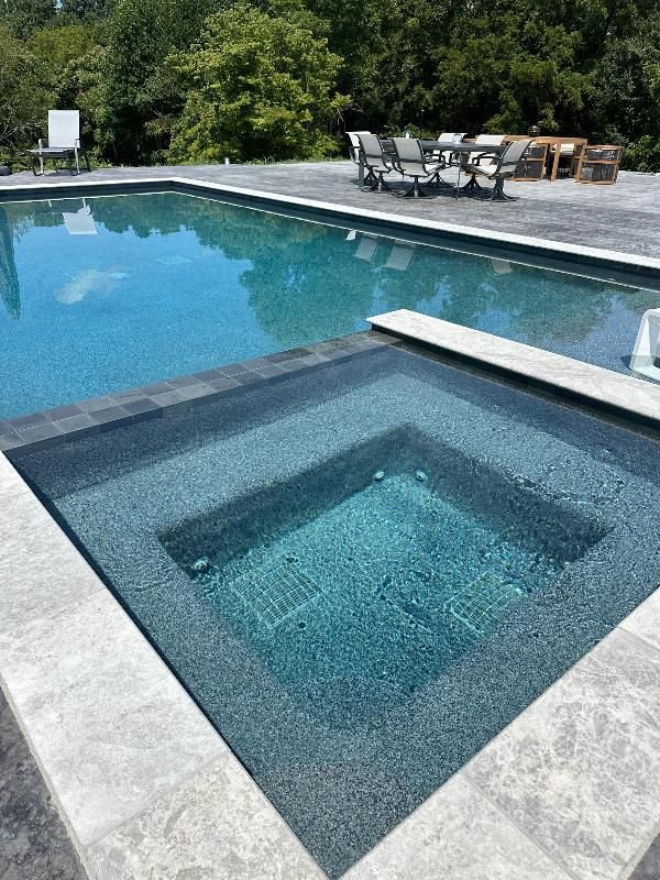 Pool with attached spa, both filled with teal water, on a grey stone patio. Furniture visible in the background.