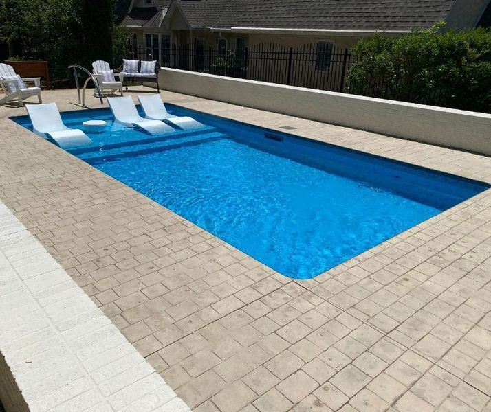Rectangular blue pool with white in-pool chaise lounges, surrounded by light concrete. A house is in the background.
