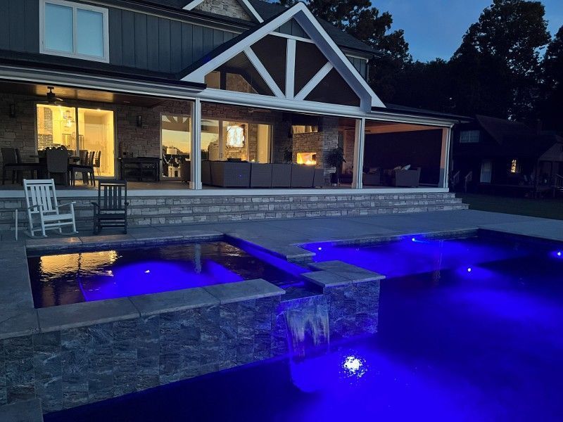 Blue swimming pool with built-in steps and connected hot tub, surrounded by brick patio.