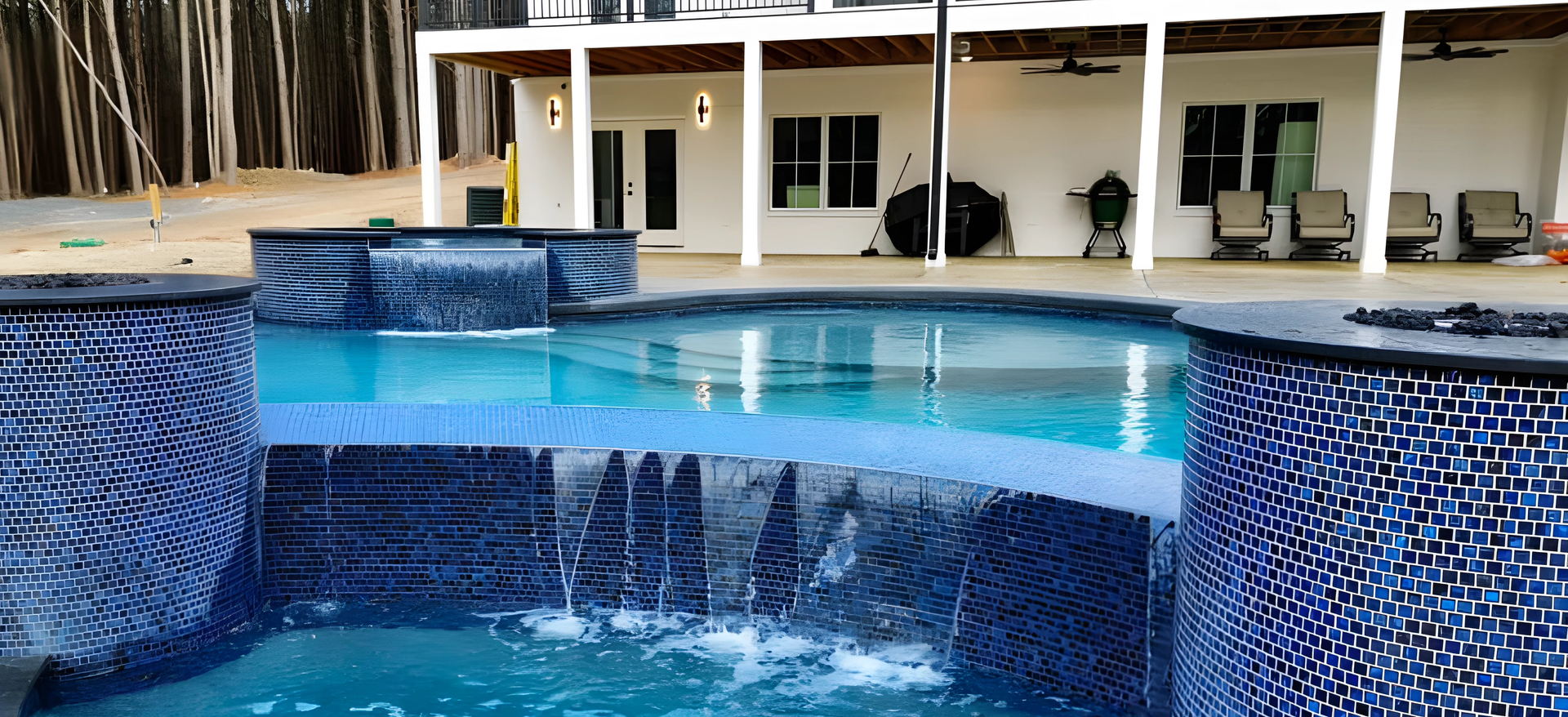 Swimming pool with blue tile and waterfall feature, in front of a white house with a covered patio.