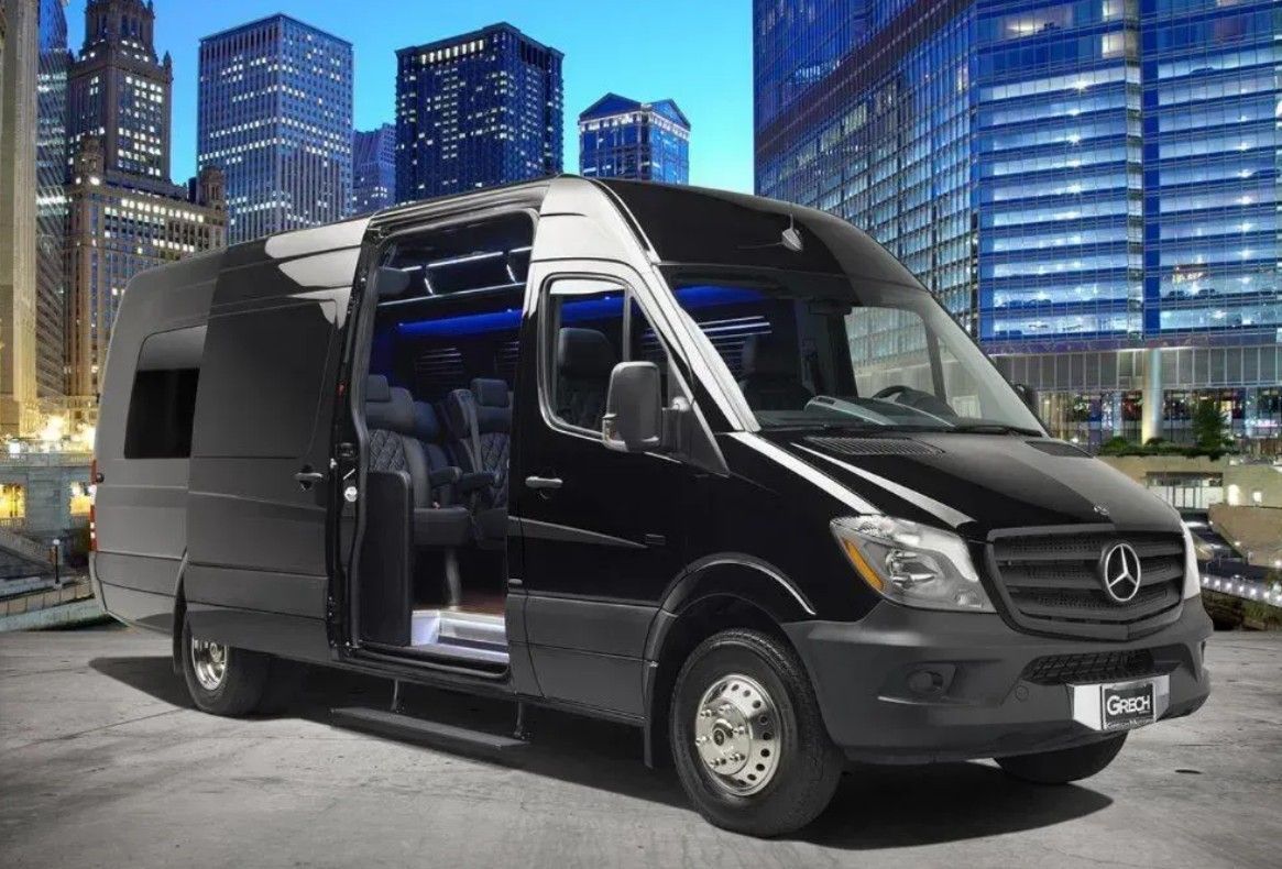Black Mercedes Sprinter van with open door parked in front of a city skyline at night.
