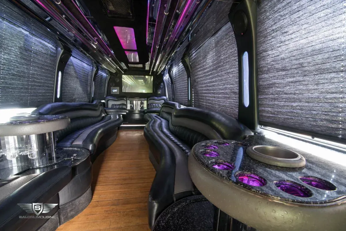 Interior of a black luxury party bus with seating, tables, and a TV. Pink and purple lighting accents.