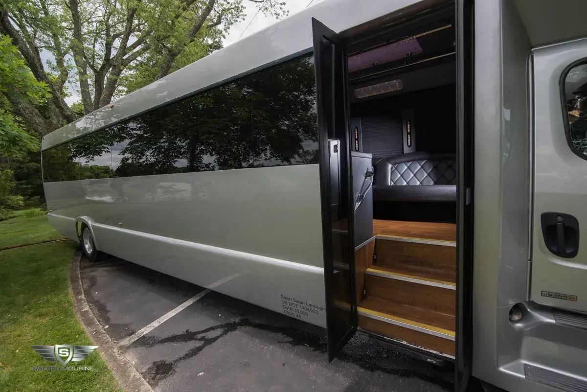 Silver party bus with open door revealing interior, parked on asphalt beside grass.