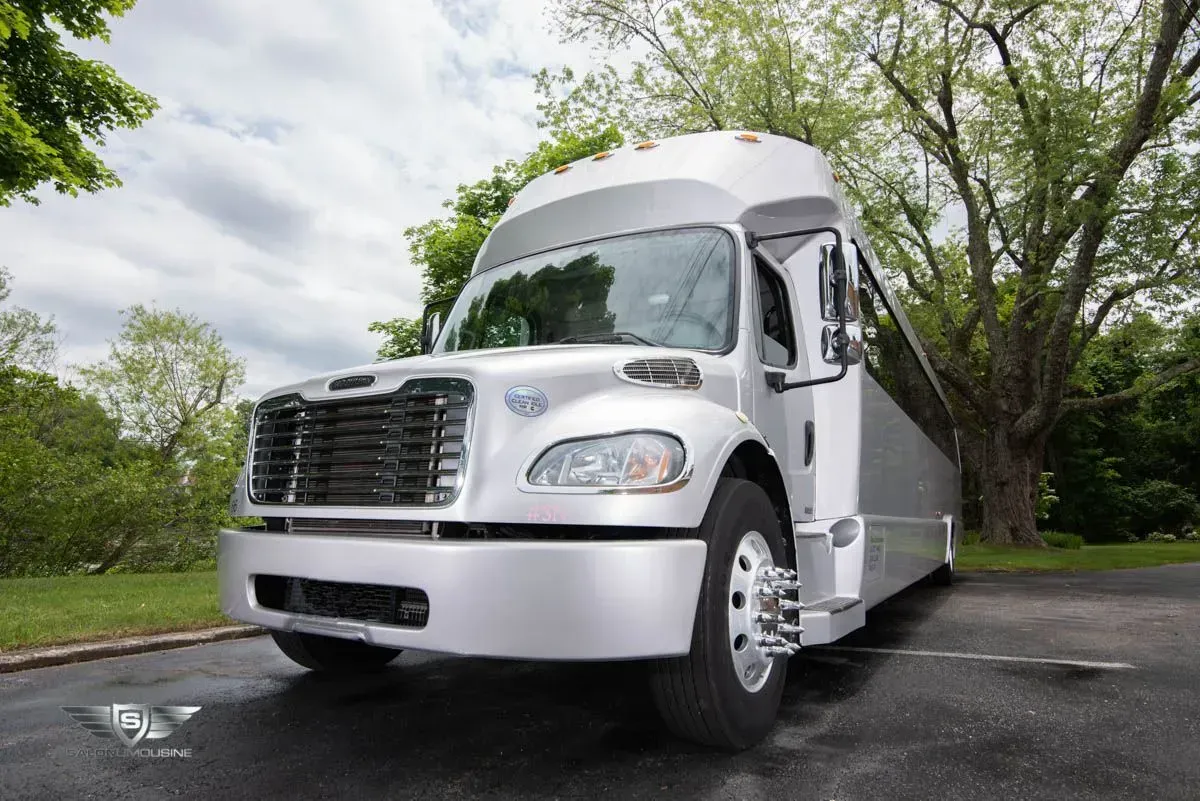 Silver passenger bus parked on asphalt, cloudy sky, trees in the background.
