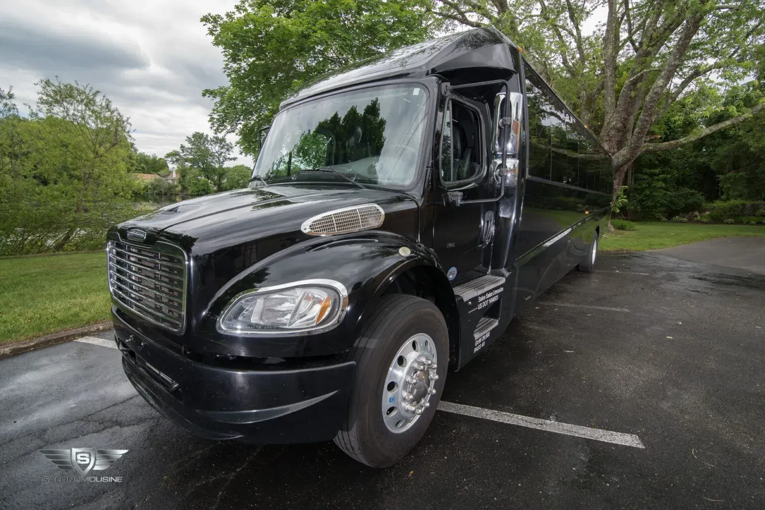 Black bus parked on wet pavement, surrounded by greenery under a cloudy sky.