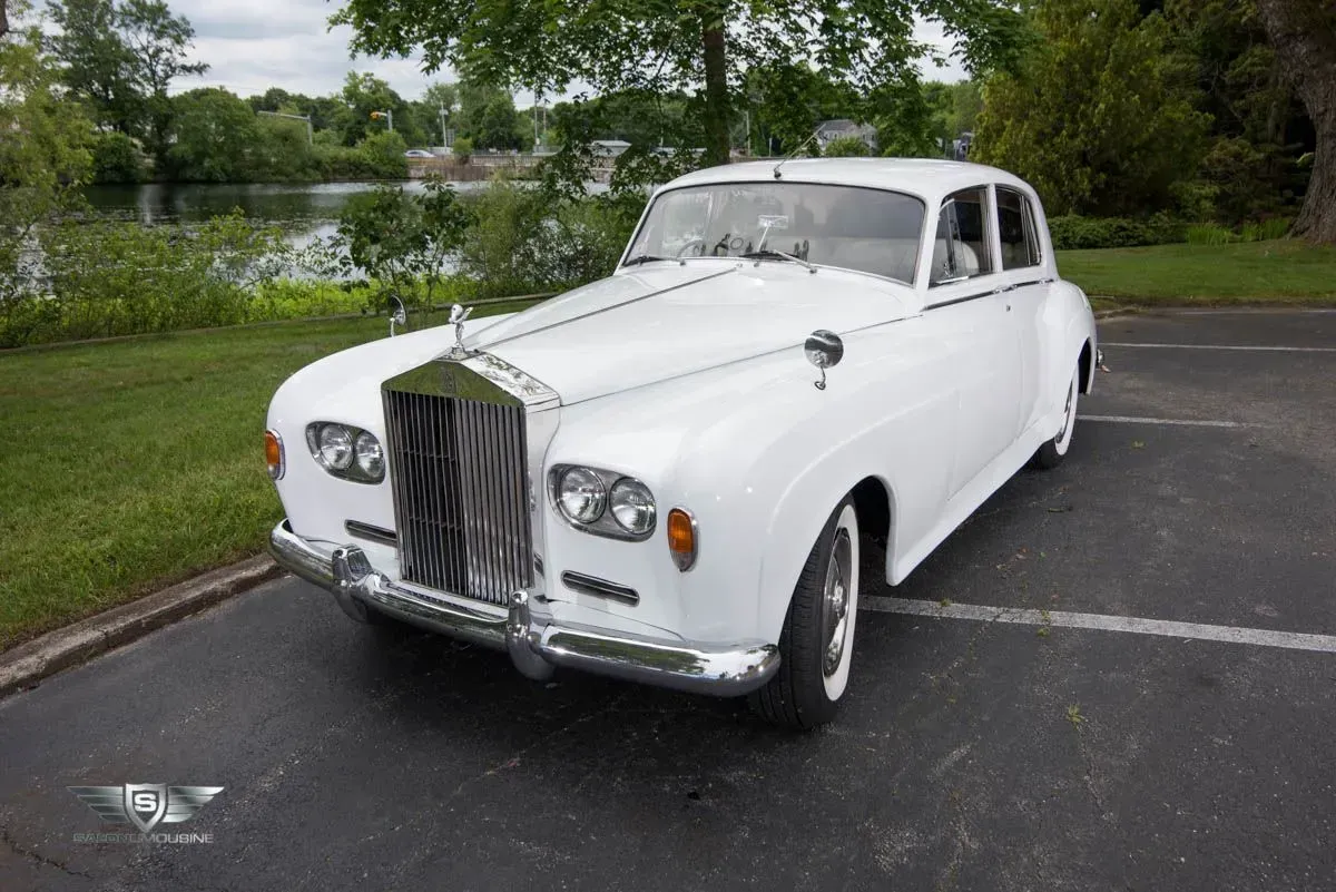 White vintage Rolls Royce sedan parked near a lake, on a paved surface.