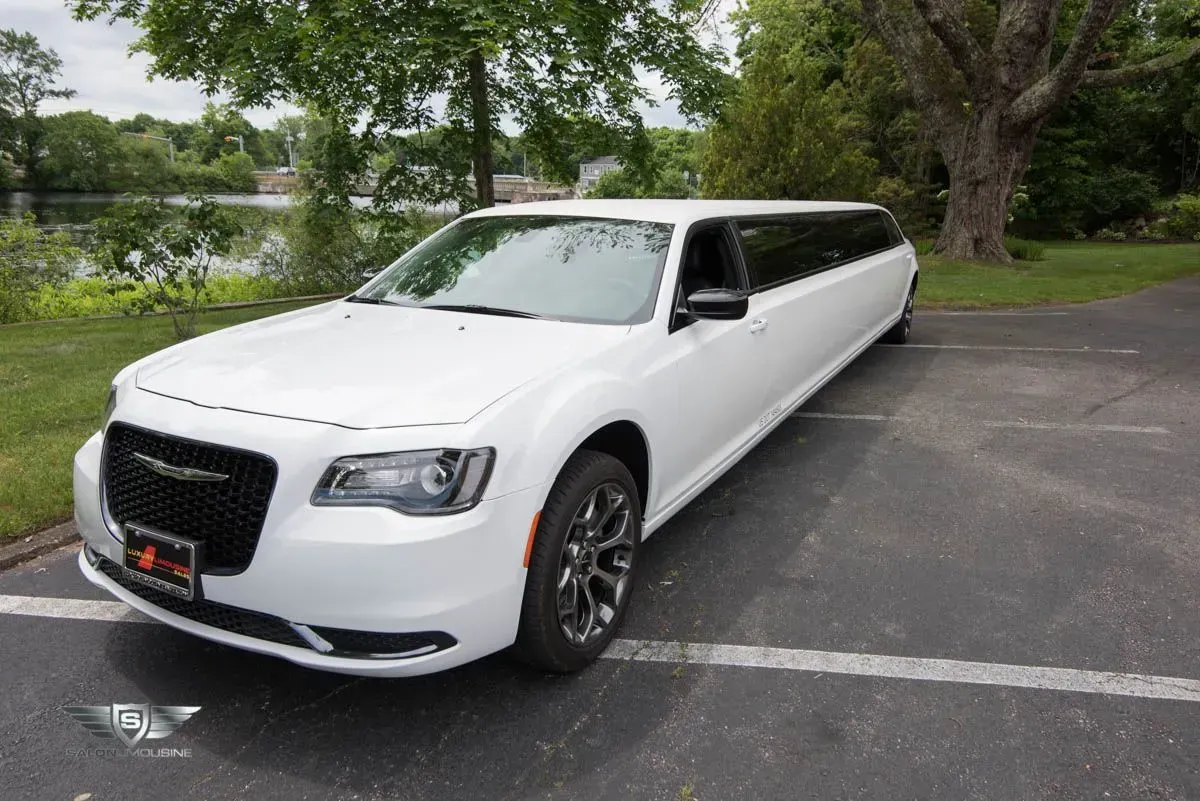 White Chrysler 300 limousine parked on a paved lot near a lake.
