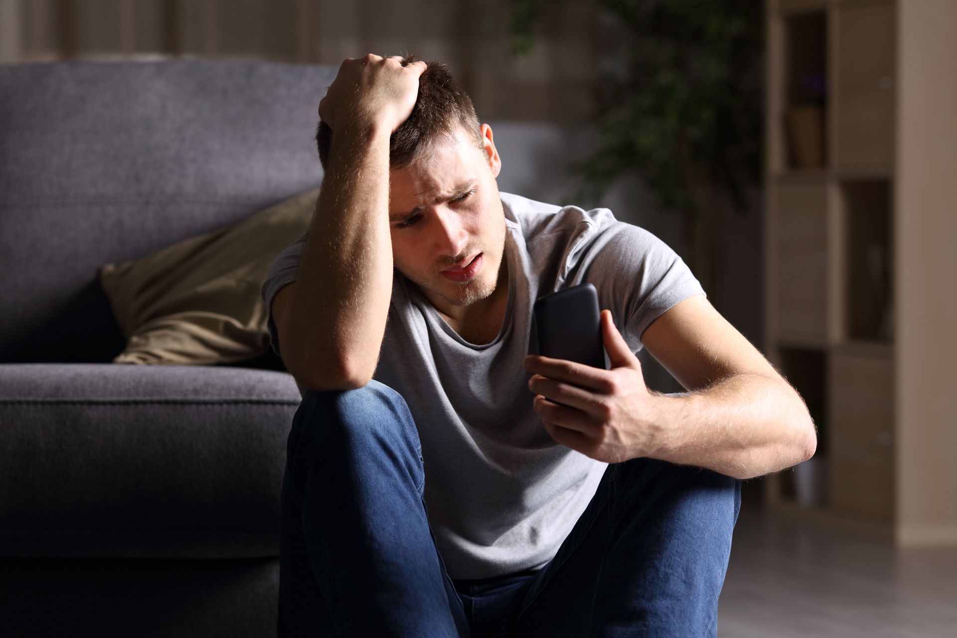 Man sitting on the floor by a couch, looking at his phone with a worried expression