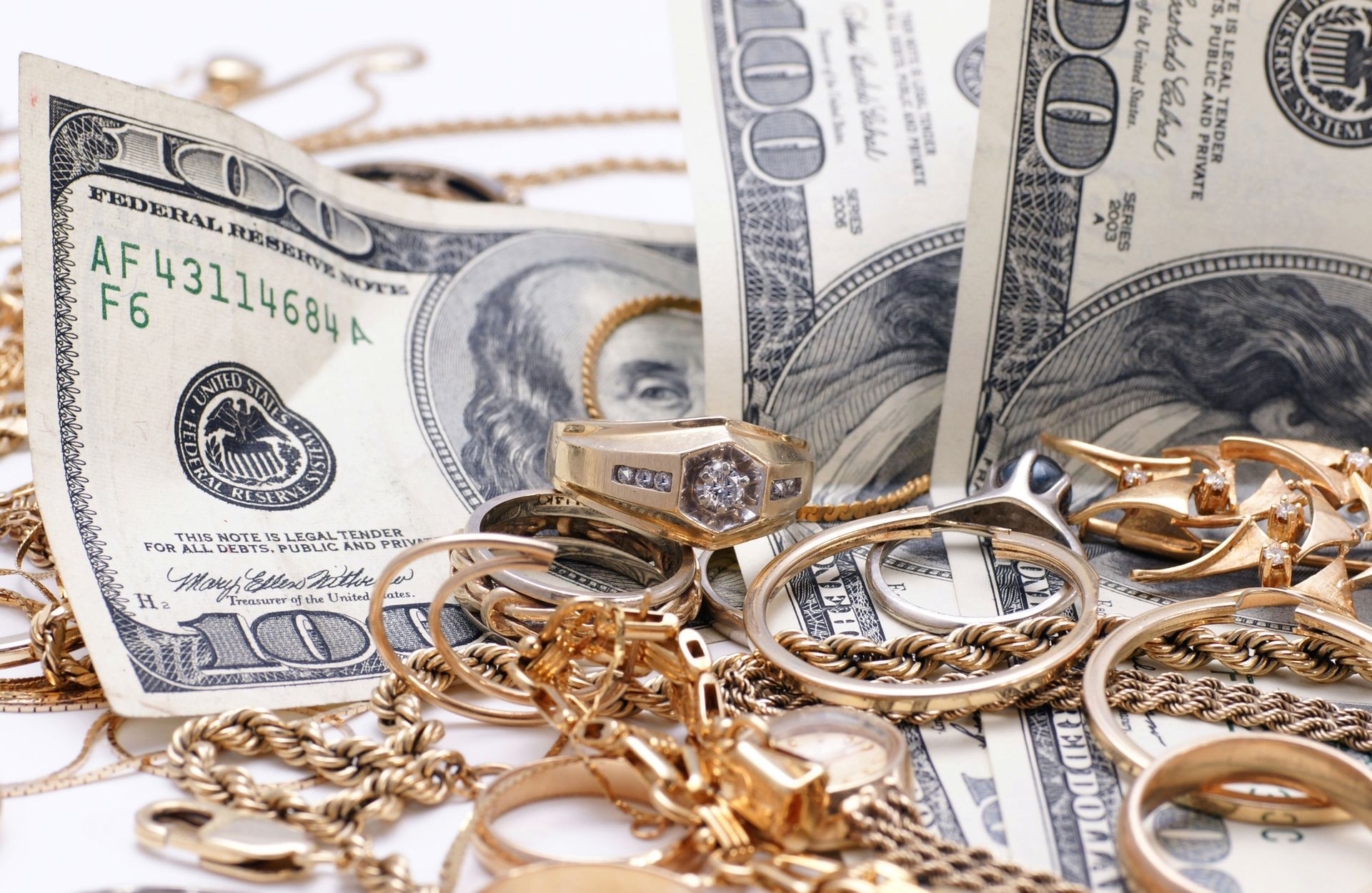 Gold jewelry and US dollar bills arranged in a pile on a white surface