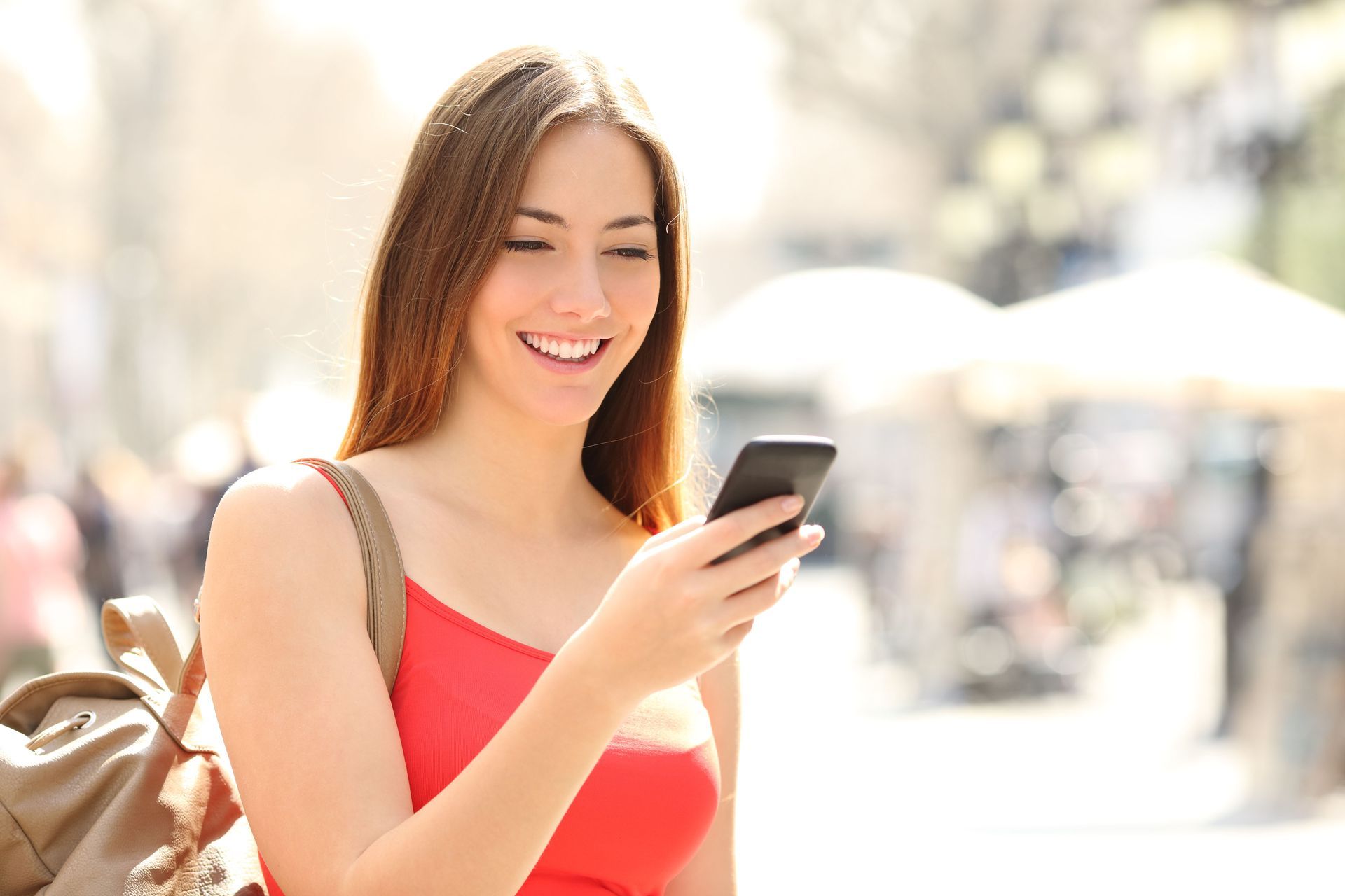 Smiling woman in a red top looking at her phone outdoors, carrying a backpack
