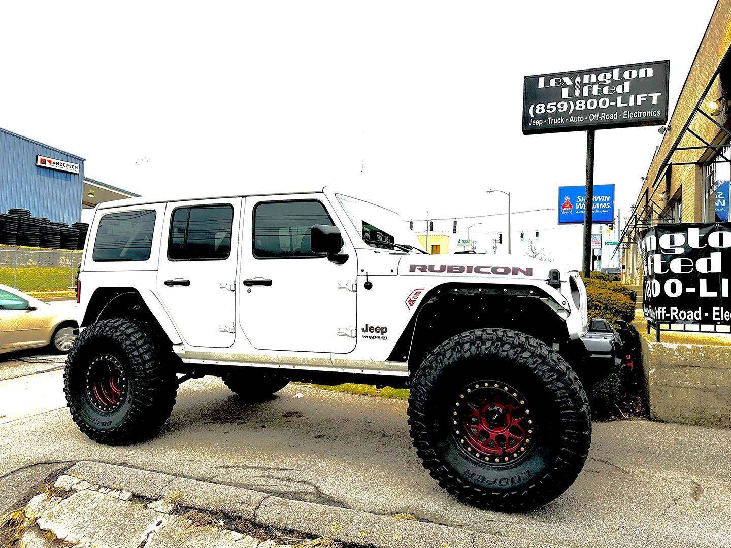 White lifted Jeep Wrangler parked outside a shop, with black off-road tires and red rims