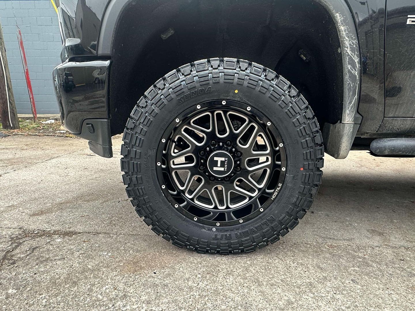 Black pickup truck wheel with rugged off-road tire and black alloy rim on pavement