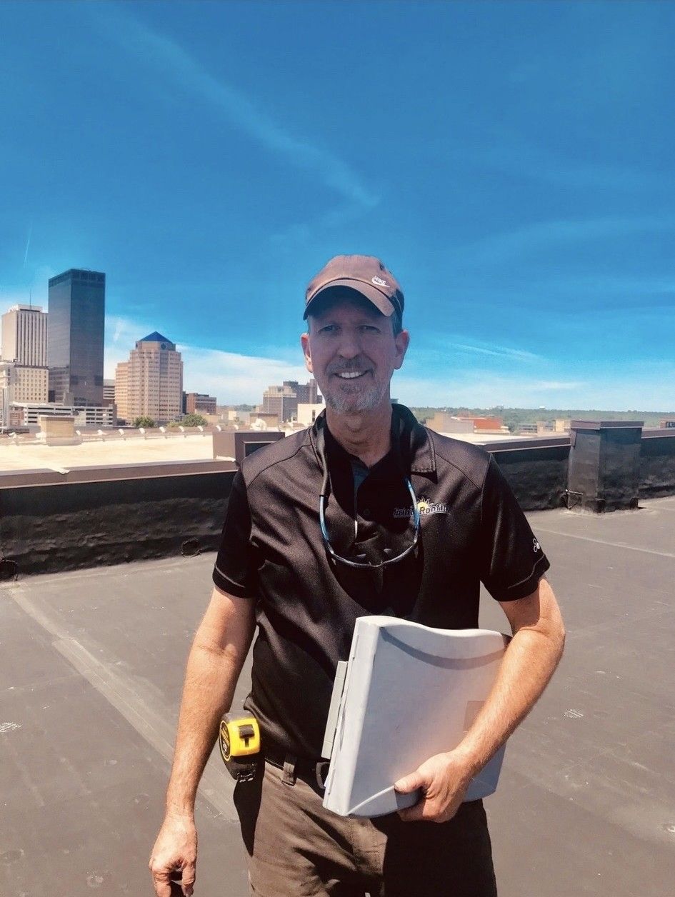 Man on rooftop with city skyline, holding a gray device. Wearing a hat, smiling, and wearing a black shirt.