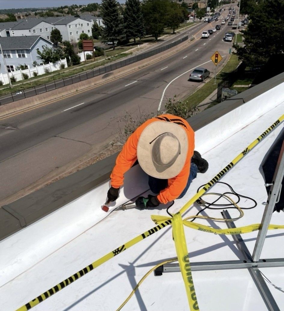 Roofer in orange shirt and straw hat, painting white roof near a busy road, with safety tape.