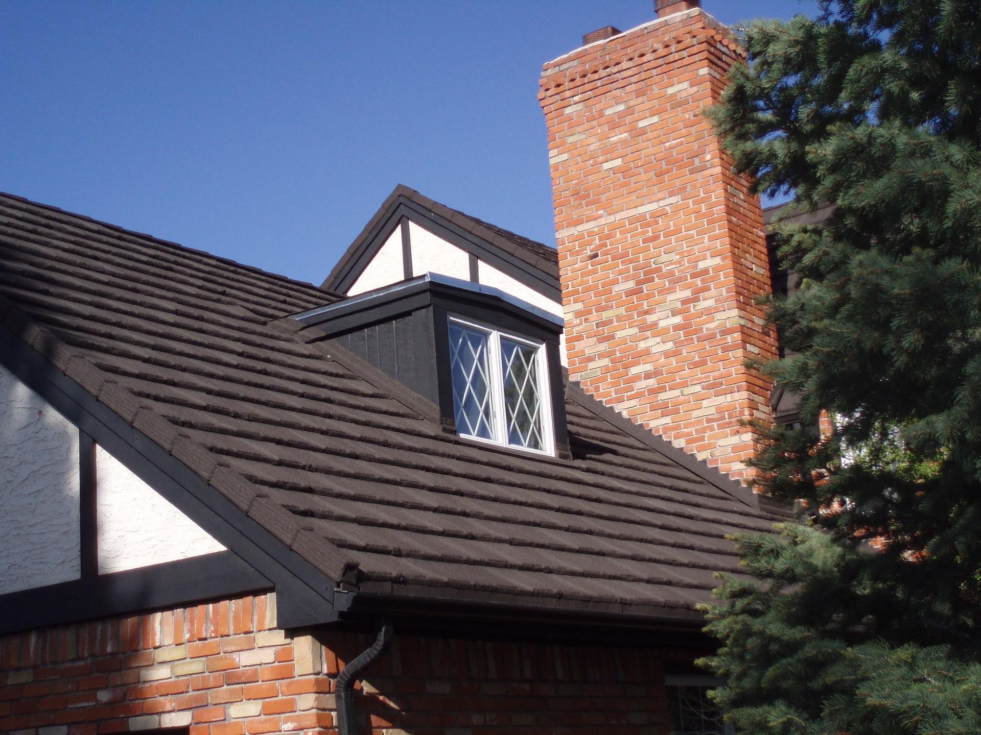Brown roof of a brick house with a dormer, tall brick chimney, and evergreen tree against a blue sky.