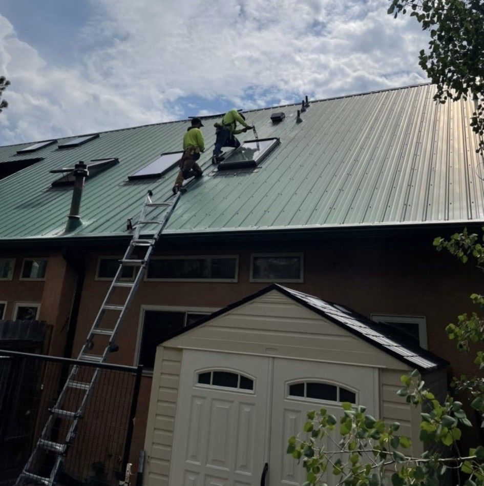 Two workers installing solar panels on a green metal roof, ladder against the building.